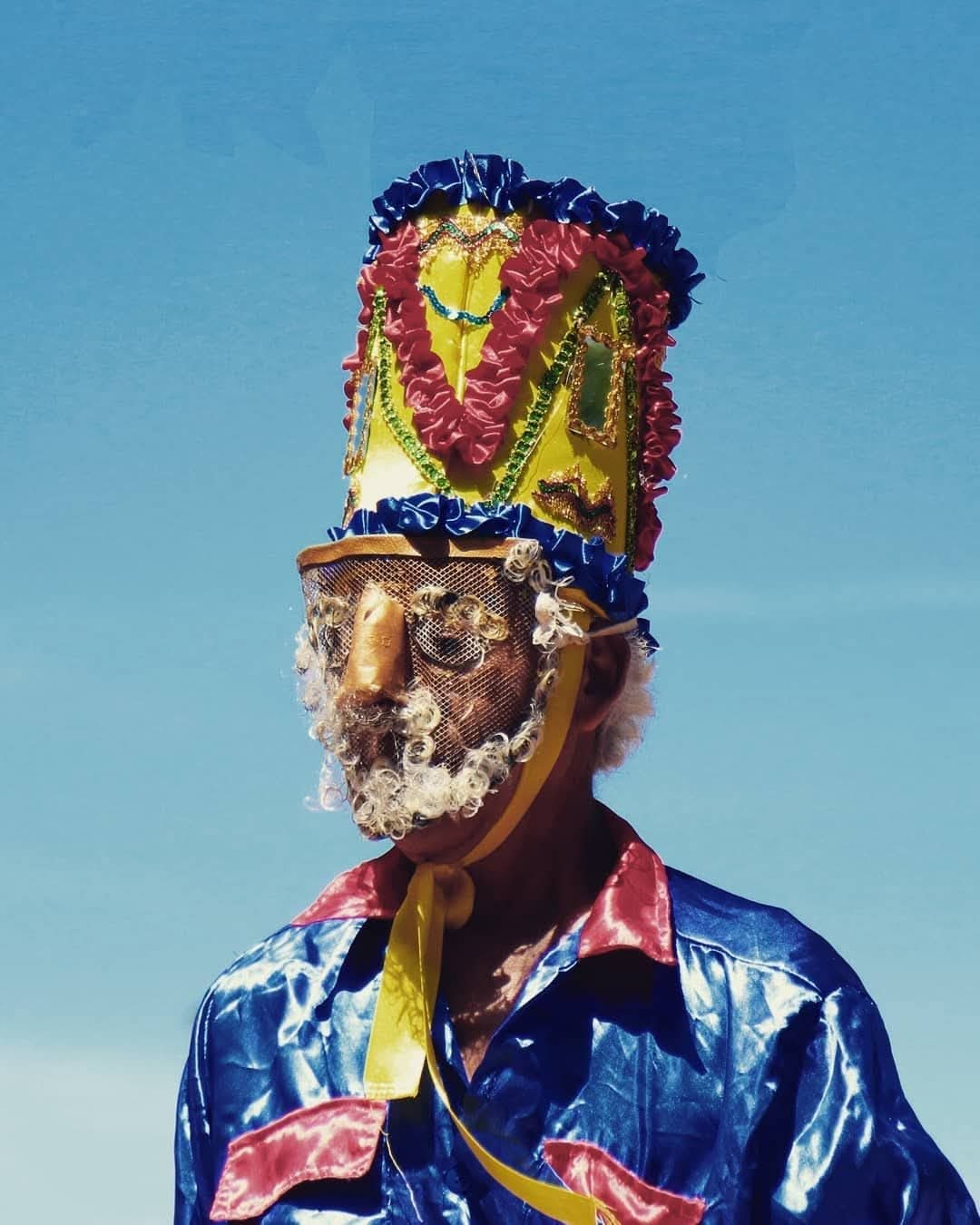 Personaje del sainete con máscara, gorro adornado y traje de raso, contra el cielo